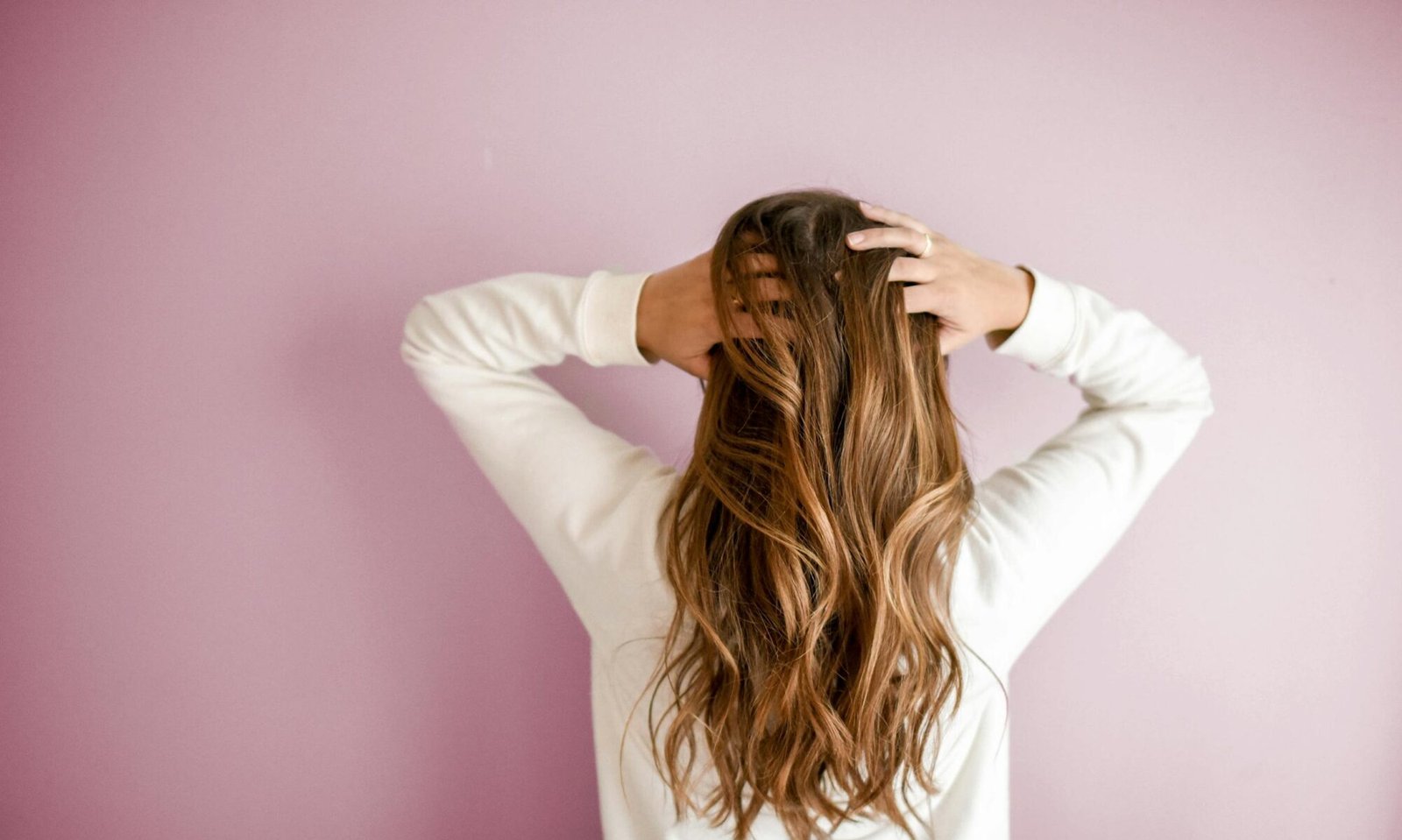 Back view of a woman with elegant long brown hair against a pink wall, showcasing stylish hair design.